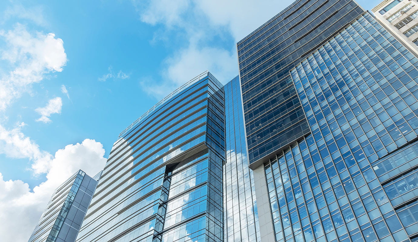 A modern cityscape featuring several tall glass-structured office buildings against a blue sky with scattered clouds.