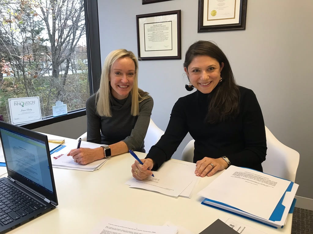 Jessica Bailey and Ali Cooley sit at a desk and smile for the camera